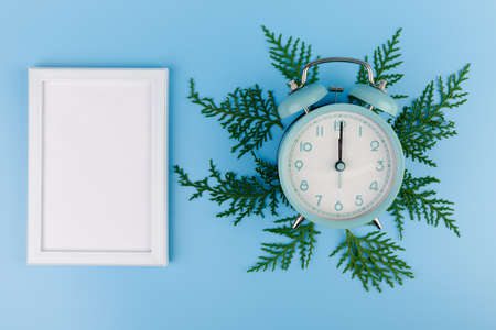 Blank white photo frame, blue alarm clock and green cypress branch on blue background. Christmas composition, copy space.の写真素材