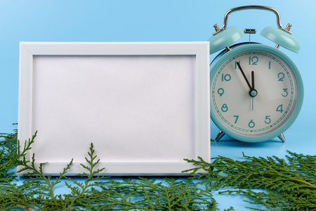 Blank white photo frame, blue alarm clock and green cypress branch on blue background. Christmas...