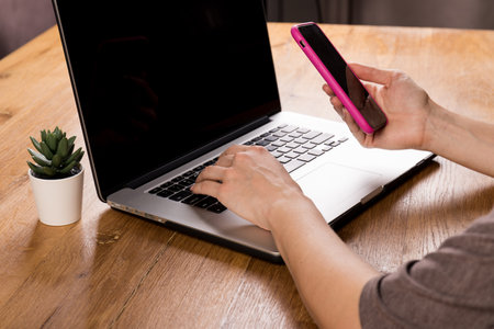Cropped image of a woman holding a smartphone to receive a message confirming the transaction on a laptop, a woman uses a mobile application to synchronize data with a netbook via bluetoothの写真素材