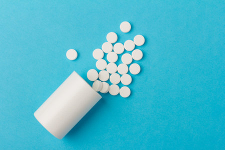 Medication bottle and white pills spilled on blue pastel coloured background. Medication and...