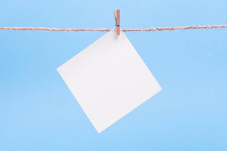A white square piece of note paper hangs on clothespins on a blue background, mock upの写真素材