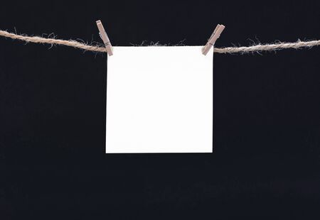 A white square piece of note paper hangs on clothespins on a black background, mock upの写真素材