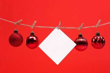 A white square piece of note paper hangs on clothespins on a red background whith Christmas ball , mock upの写真素材