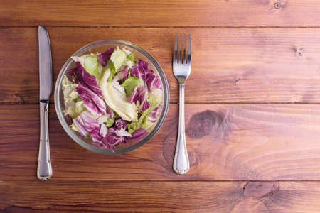 Fresh vegetable salad leaves in a glass bowl with a fork and knife, isolated on a wooden background close-upの写真素材