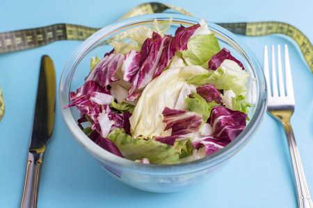Fresh vegetable salad leaves in a glass bowl with a fork and knife, isolated on a blue background close-upの写真素材