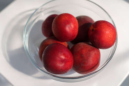 Ripe red plum-peach, nectarine in a glass container on a white background with a play of shadowsの写真素材