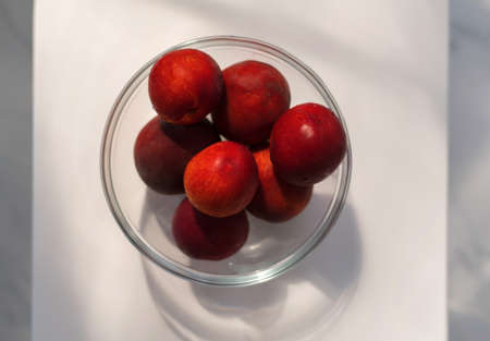 Ripe red plum-peach, nectarine in a glass container on a white round background with a play of shadowsの写真素材