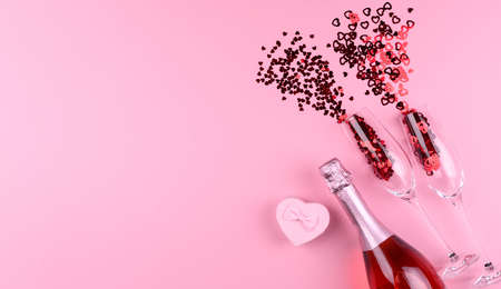 Two champagne glasses filled with heart shaped sequins with a bottle of champagne and a Valentine's Day gift on a pink background.の写真素材