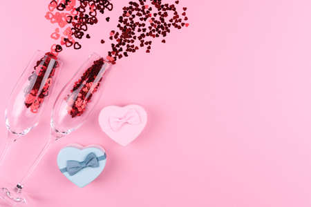 Two champagne glasses filled with heart shaped sequins with a bottle of champagne and a Valentine's Day gift on a pink background.の写真素材