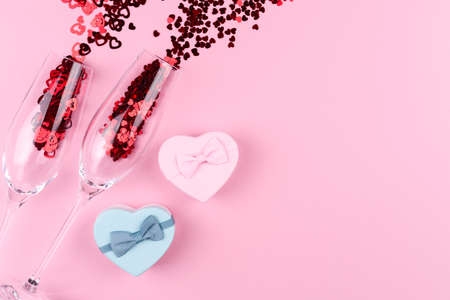 Two champagne glasses filled with heart shaped sequins with a bottle of champagne and a Valentine's Day gift on a pink background.の写真素材