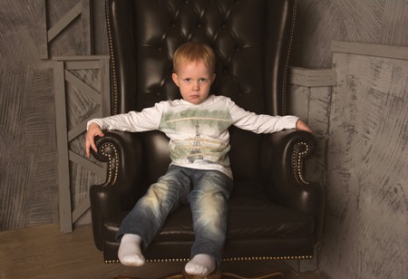 blond boy in jeans sitting in a black leather chairの写真素材