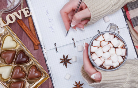Women's hands holding a pen and a cup of cocoa or hot chocolate with marshmallow on the background...