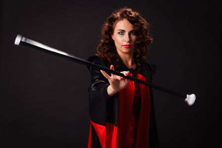 red-haired beautiful young woman in a red and black cape rotates in the fingers stick on a dark backgroundの写真素材