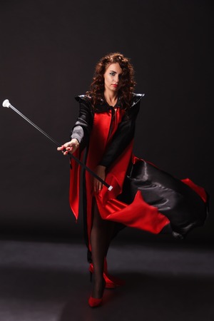 A beautiful young woman in a long fold of red and black fabric and in red suede high-heeled shoes waving the cloak of the cloak and spinning in her fingers a cane on a dark backgroundの写真素材