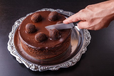 A mans hand with a kitchen knife above a chocolate homemade biscuit cake on a metal plateの写真素材