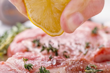 The man's hand squeezes the lemon juice with a raw pork steak. Also on the table, onion, thyme, garlic, salt, pepper, salt and pepper shaker on a wooden cutting board close-upの写真素材