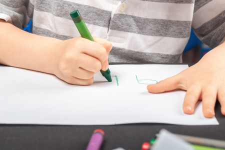 Drawing on a white sheet of paper with colored wax pencils. The concept of children's creativity and hobbies. A child draws a green pencil on white paper. Baby hands closeupの写真素材