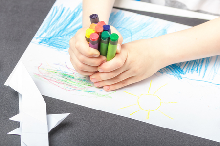 Drawing on a white sheet of paper with colored wax pencils. The concept of children's creativity and hobbies. The child draws a set of colored pencils abstract lines and patterns. Baby hands closeupの写真素材