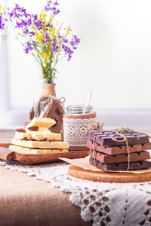 Still life with dark, milky and white chocolate bars, dishes and a bouquet of wild flowers against the window. Items are located on burlap and lace napkins.の写真素材
