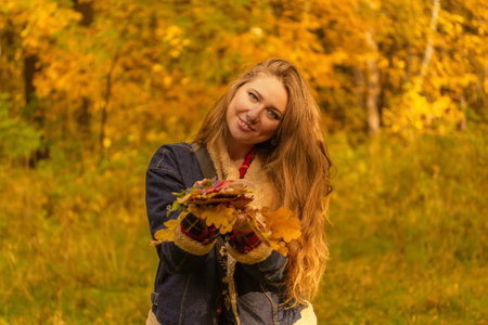Autumn concept. Young red-haired woman in autumn forest smiles and holds fall foliage. High quality...
