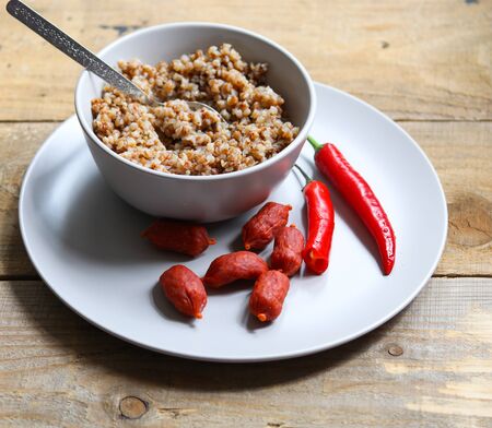 buckwheat porridge in a white bowl with sausages and chili peppers on a wooden tableの写真素材