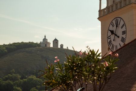 A small chapel on a hill in the backlight. In the foreground are tower clocks and flowers. Chapel of...