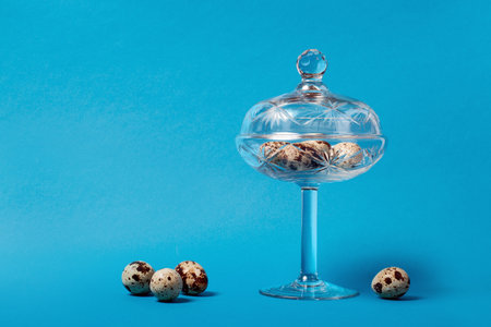 Variegated, quail eggs on the surface and in a glass, transparent vase with a high stem. Blue background. Easter.の写真素材