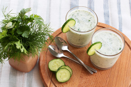 On a table on a wooden stand are two glass cups with a smoothie with cucumber and greens, a bunch of greens, metal spoons.の写真素材
