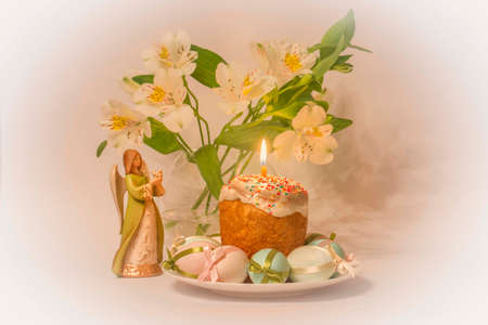 Easter. Pastry decorated with colored beads and a burning candle. Traditional colored eggs decorated with ribbons. There is an angel figurine on the table. In the background, a vase with flowers.の写真素材
