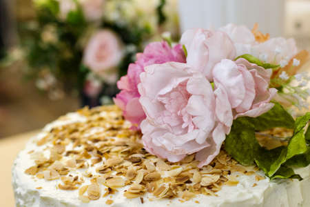 Delicious white cream cake, garnished with roasted almonds and edible pink peonies with green leaves. Banquet, celebration, confectionery. Close-up. Bokeh.の写真素材