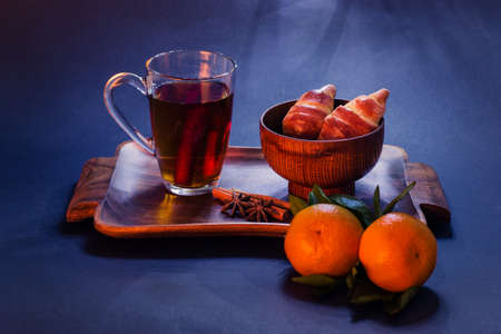 A glass, transparent cup of tea with cinnamon sticks is on a blue background of the table on a wooden tray. Buns are in the bowl. Nearby are two ripe, bright tangerines.の写真素材