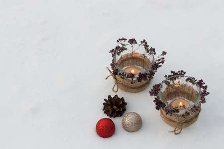 Against the background of white snow, there are two glass candlesticks, decorated with sprigs of oregano with burning candles. The Christmas composition is complemented by cones and balls. Outdoors.の写真素材