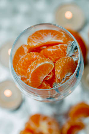 Glass, transparent goblet filled with bright mandarin slices. Candles are burning on a white fabric background, bokeh. Festive, Christmas decoration. View from above.の写真素材