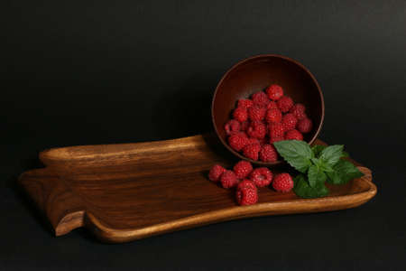 Ripe red raspberries with a green leaf spilled out of a wooden bowl onto a tray. Black background. Close-up.の写真素材