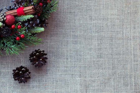 Background from natural fabric. In the corner is a part of a Christmas wreath with green branches, red berries, cinnamon sticks, Christmas balls. Two fir cones. Place for an inscription. Close-up.の写真素材
