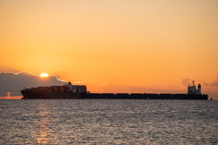 Dramatic sunrise casting reflections on rippling water and mountains, Container vessel bathed in dawn light with shimmering water reflection and scenic backdropの写真素材