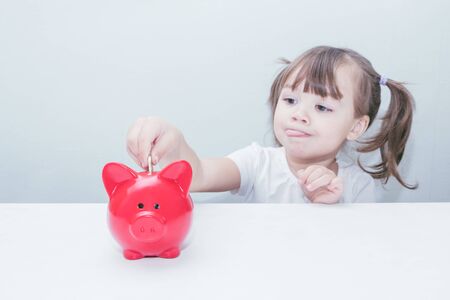 child girl dips a coin in a red piggy bank standing on the tableの写真素材