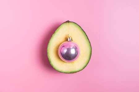 Green ripe avocado with a pink Christmas ball instead of a stone on a pink background. Creative concept of healthy food for New Years tableの写真素材