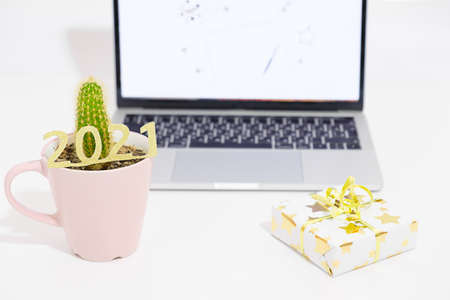 Office cactus in pink cup decorated with 2021 golden numbers. New Years gift box in front of laptop