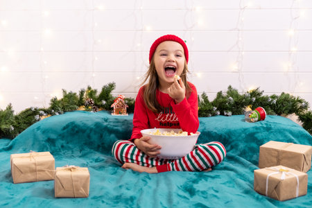 little girl in New Years costume eats popcorn from white bowl while sitting on the bed. Traditional holiday decorations on Christmasの写真素材
