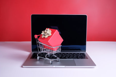 A shopping cart with a red gift box stands on a laptop keyboard. A laptop on a white table against a red wall surrounded by decorations. Online gift shopping conceptの写真素材