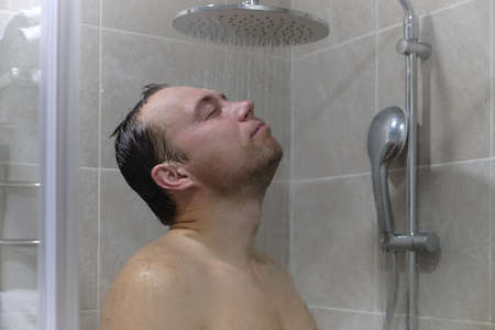 young man takes a shower. he stands in a shower stall with his face under the jets of tropical rain...