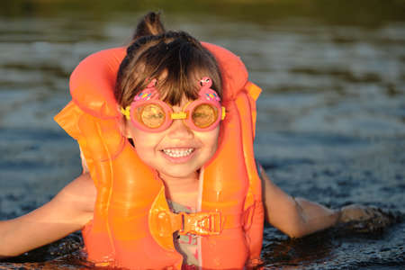A little girl bathes in the water in the sunset light. Child playing on the beach during summer vacation. The child swims in a life jacket and goggles. Safety on the water.の写真素材