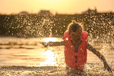 Defocused image for background. Little girl plays in the river lake. Golden hour. A child splashes water on the beach during summer vacation. Safety on the water.の写真素材