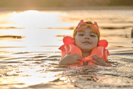 A little girl bathes in the water in the sunset light. Child playing on the beach during summer vacation. The child swims in a life jacket and goggles. Safety on the water.の写真素材