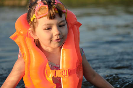 A little girl bathes in the water in the sunset light. Child playing on the beach during summer vacation. The child swims in a life jacket and goggles. Safety on the water.の写真素材