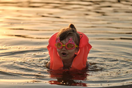 A little girl bathes in the water in the sunset light. Child playing on the beach during summer vacation. The child swims in a life jacket and goggles. Safety on the water.の写真素材