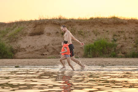 Daughter with her father plays in the river lake. A child in a life jacket. Safety on the water. Fun time during family vacations. Golden hour.の写真素材