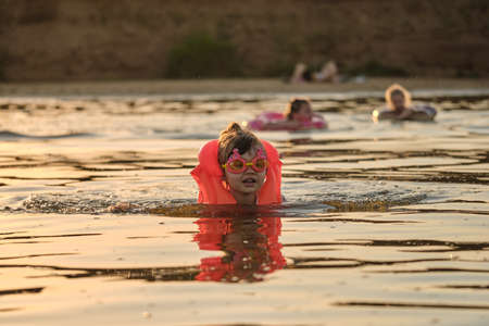 A little girl bathes in the water in the sunset light. Child playing on the beach during summer vacation. The child swims in a life jacket and goggles. Safety on the water.の写真素材