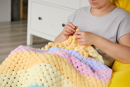 A young woman knits a warm bright plaid from multi-colored terry looped yarn with her hands. A cozy pastime for a hobbyの写真素材